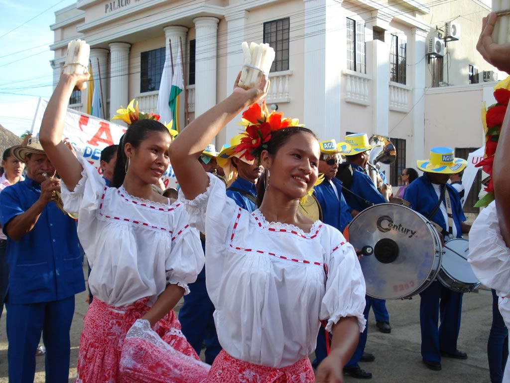 Dos jóvenes con blusas blancas y flores rojas en el pelo bailan cumbia sosteniendo en alto manojos de velas, ante una banda de músicos.